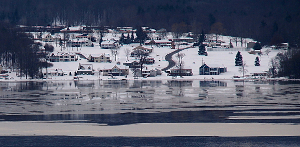 [Along the snow-covered hillside leading down to the lake are several dozen two and three-level houses. The lake in the foreground was iced over but patches have begun to melt.]